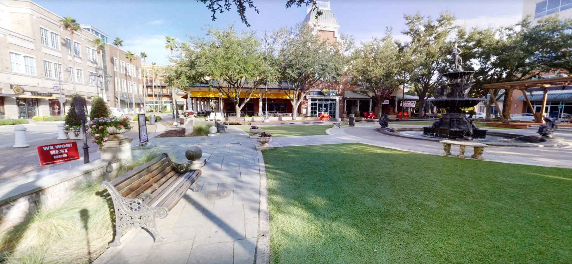 Sunny outdoor shopping plaza with a central lawn, stone fountain, and storefronts under a blue sky